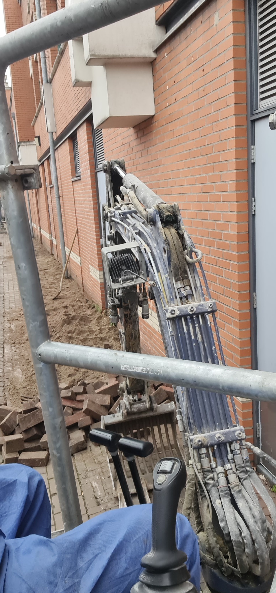 Mini koparka przy ceglanym budynku, widoczne porozrzucane cegły i ziemia, fragment rusztowania w kadrze, operator w niebieskim ubraniu.