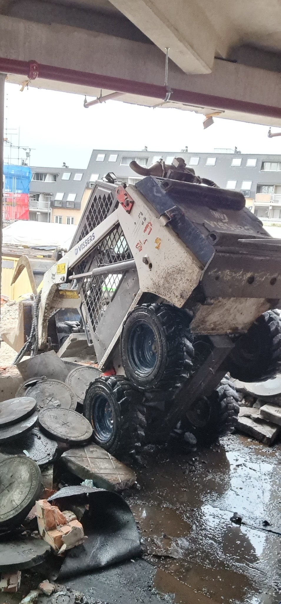 Miniładowarka Vissers po częściowym zawaleniu ściany w Częstochowie, widoczne gruzowisko i mokra posadzka, w tle budynki mieszkalne.