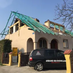 Dom w trakcie przebudowy dachu, widoczna konstrukcja drewniana i zielone elementy, na podjeździe zaparkowany samochód z reklamą usług budowlanych.