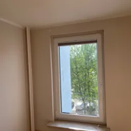 Jasne, świeżo pomalowane wnętrze z oknem wychodzącym na zieleń. Grzejnik pod oknem i rury pionowe w rogu. Neutralna kolorystyka ścian.