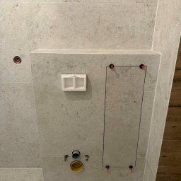 Marceli Morzecki - Ściana w łazience wyłożona beżowymi płytkami imitującymi kamień. Widoczne otwory na instalacje, rewizja i elementy montażowe. Użyto poziomowania płytek.