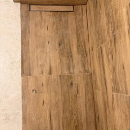 Marceli Morzecki - Ukończona kabina prysznicowa z odpływem liniowym i płytkami imitującymi drewno na ścianach i podłodze. Obok jasne, kwadratowe płytki podłogowe.