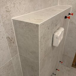 Marceli Morzecki - Obudowa WC w łazience wykończona płytkami imitującymi kamień, z widocznymi klipsami poziomującymi i fugą. Minimalistyczny design, neutralna kolorystyka.