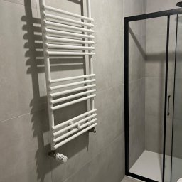GlazurnikWarszawski - Minimalistyczna łazienka z grzejnikiem drabinkowym i kabiną prysznicową z czarną ramą. Szare płytki na ścianach i podłodze. Nowoczesny design.
