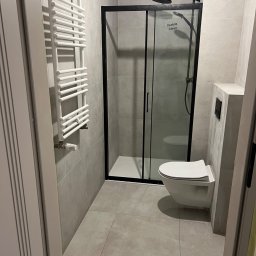 GlazurnikWarszawski - Nowoczesna łazienka w Józefowie z kabiną prysznicową w czarnej ramie, wiszącą toaletą i grzejnikiem drabinkowym. Stonowana kolorystyka płytek.