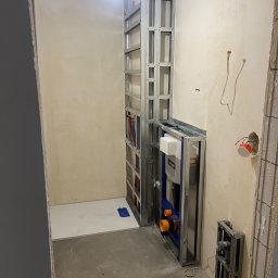GlazurnikWarszawski - Stelaż podtynkowy WC w trakcie montażu w surowej łazience. Widoczne elementy instalacji wodno-kanalizacyjnej i elektrycznej. Biały brodzik w rogu pomieszczenia.