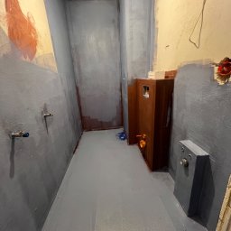 GlazurnikWarszawski - Łazienka w trakcie malowania na szaro, widoczne rury, instalacja elektryczna i zabudowa. Ściany częściowo pomalowane, sufit biały, podłoga szara.