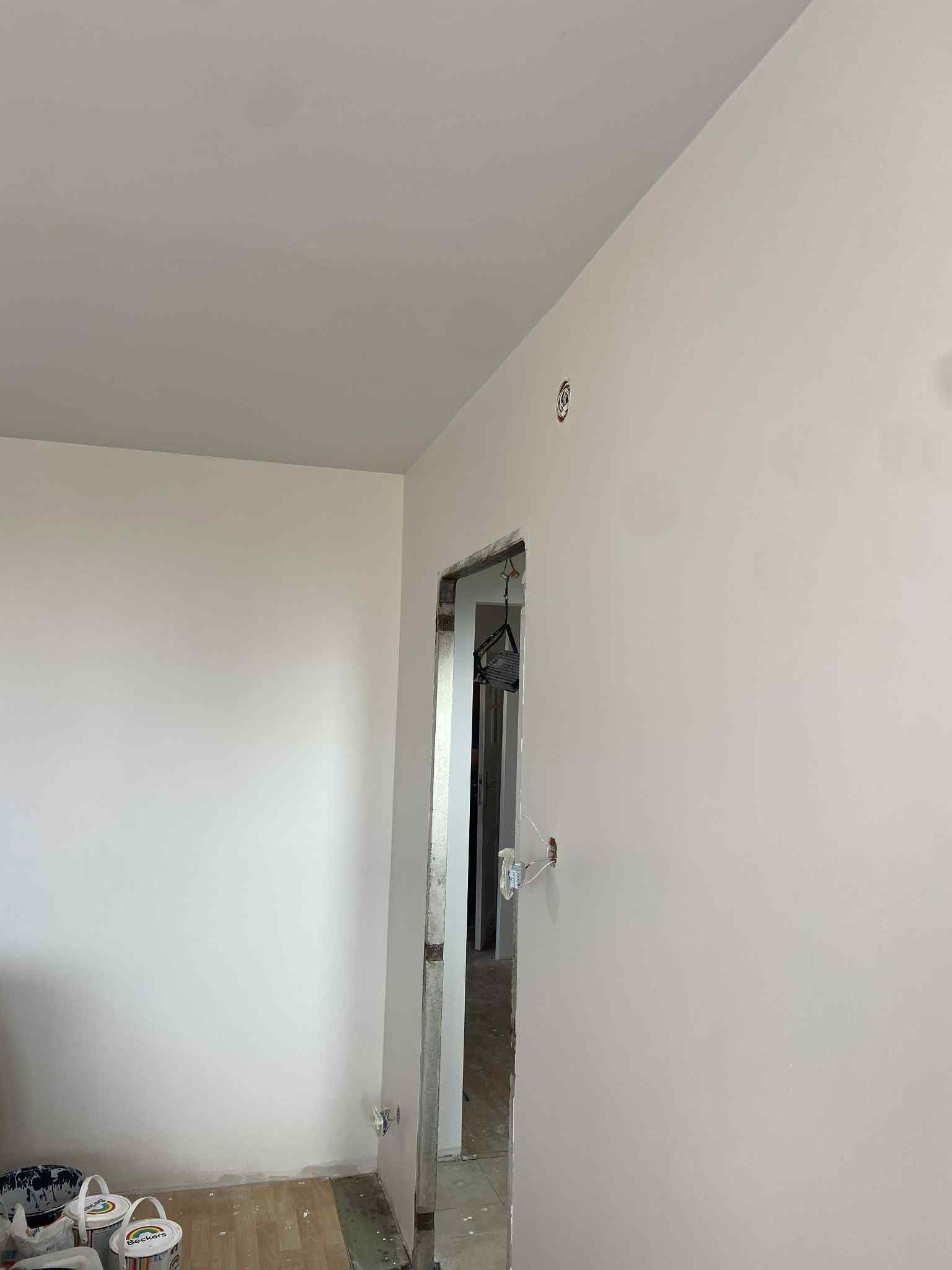 Ściana po malowaniu na jasny odcień, widoczne puszki farby Beckers na podłodze, surowa futryna drzwiowa. Widok na wnętrze pokoju.