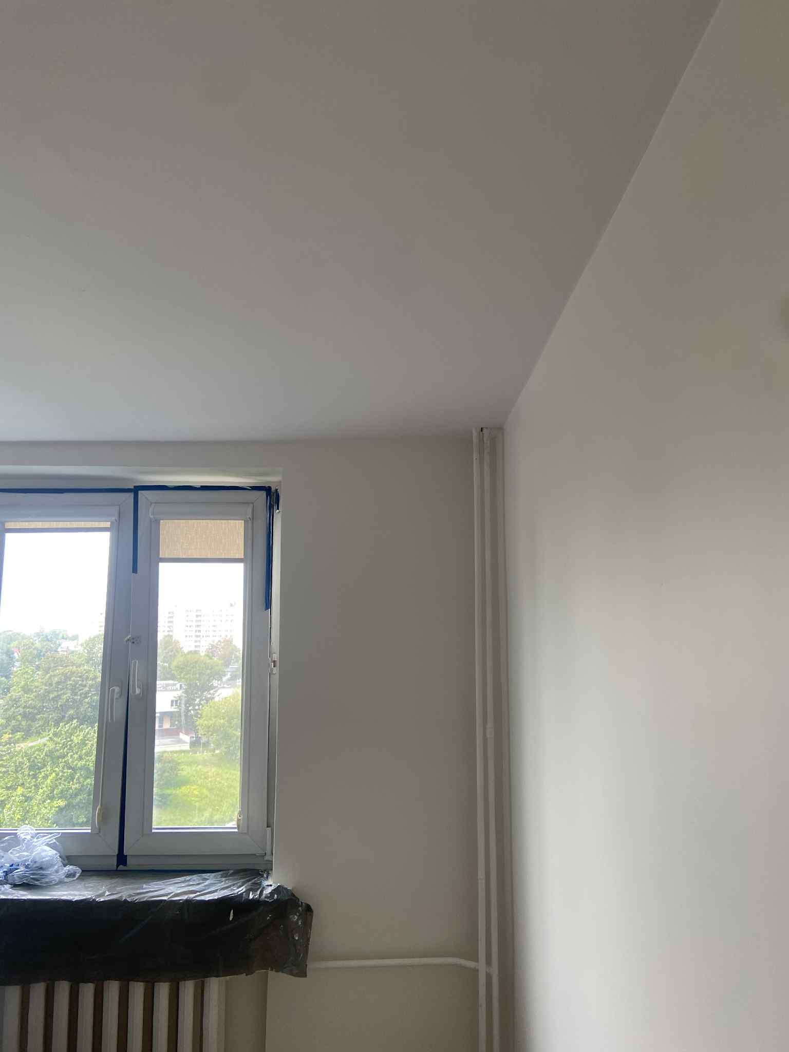 Świeżo pomalowane na biało ściany w pokoju z oknem zabezpieczonym taśmą malarską, grzejnik zakryty folią ochronną. Widok na zieleń za oknem.