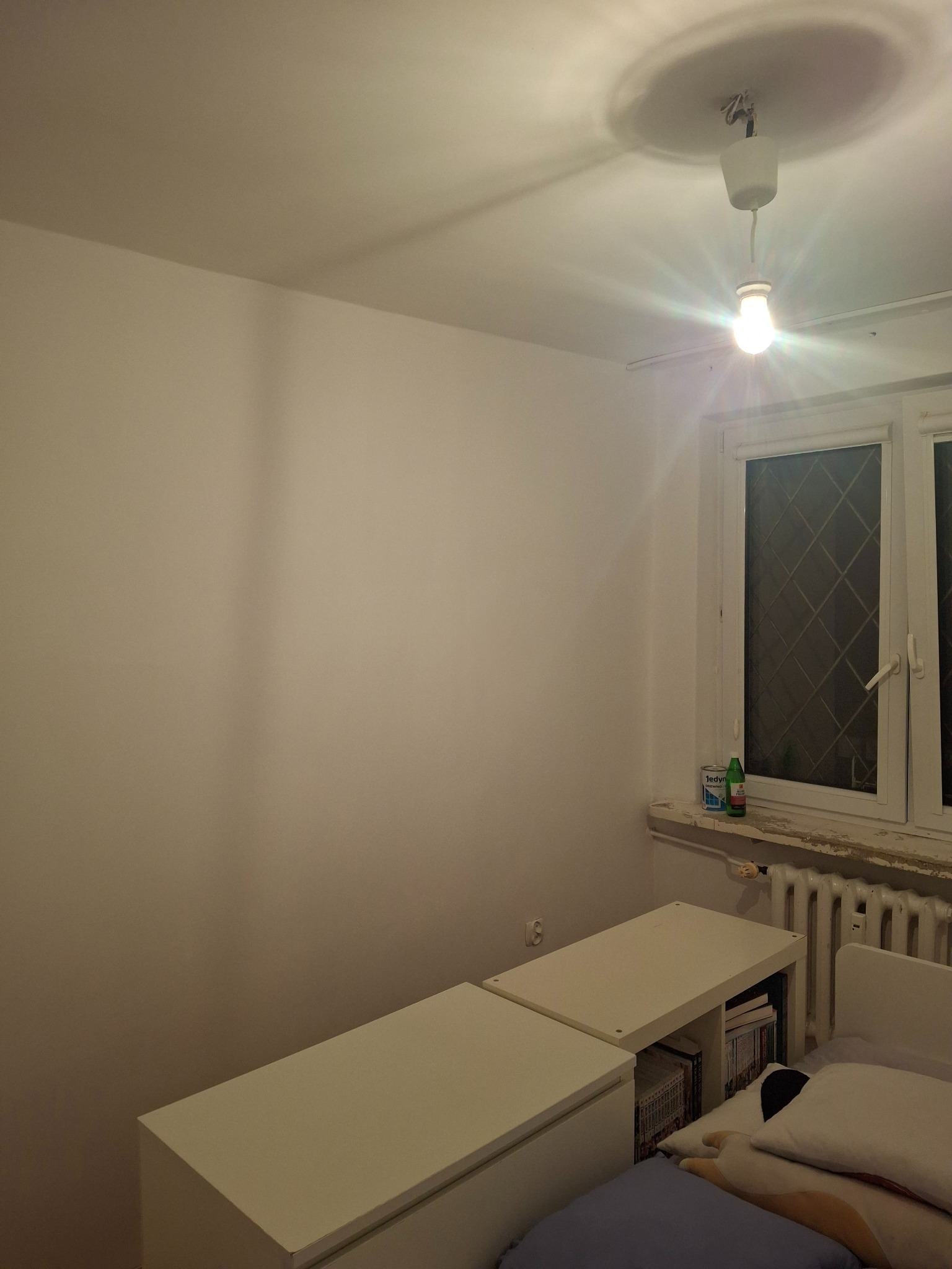 Pomalowana na gładko ściana pokoju z widoczną lampą sufitową i oknem. Na parapecie stoją puszki. Widoczny fragment biurka i łóżka.