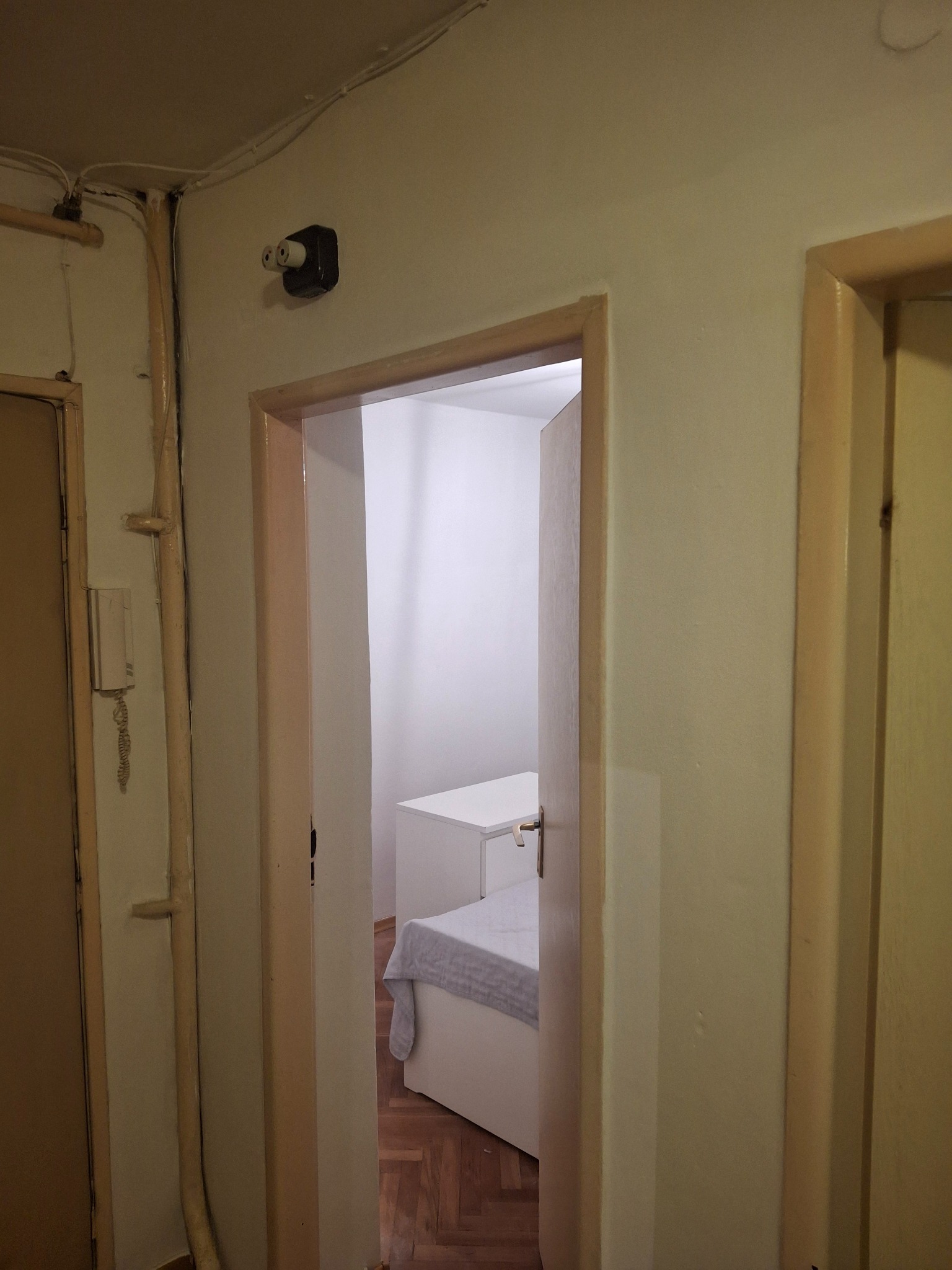 Wnętrze pokoju po malowaniu: białe ściany, widoczny fragment łóżka i biurka. Widok z korytarza przez otwarte drzwi. Stare instalacje elektryczne i rury grzewcze.