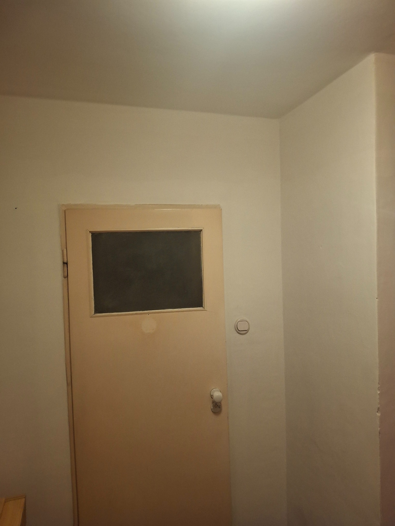 Beżowe drzwi z szybką i biała ściana z włącznikiem, widoczne ślady niedokładnego malowania. Ujęcie w rogu pomieszczenia.