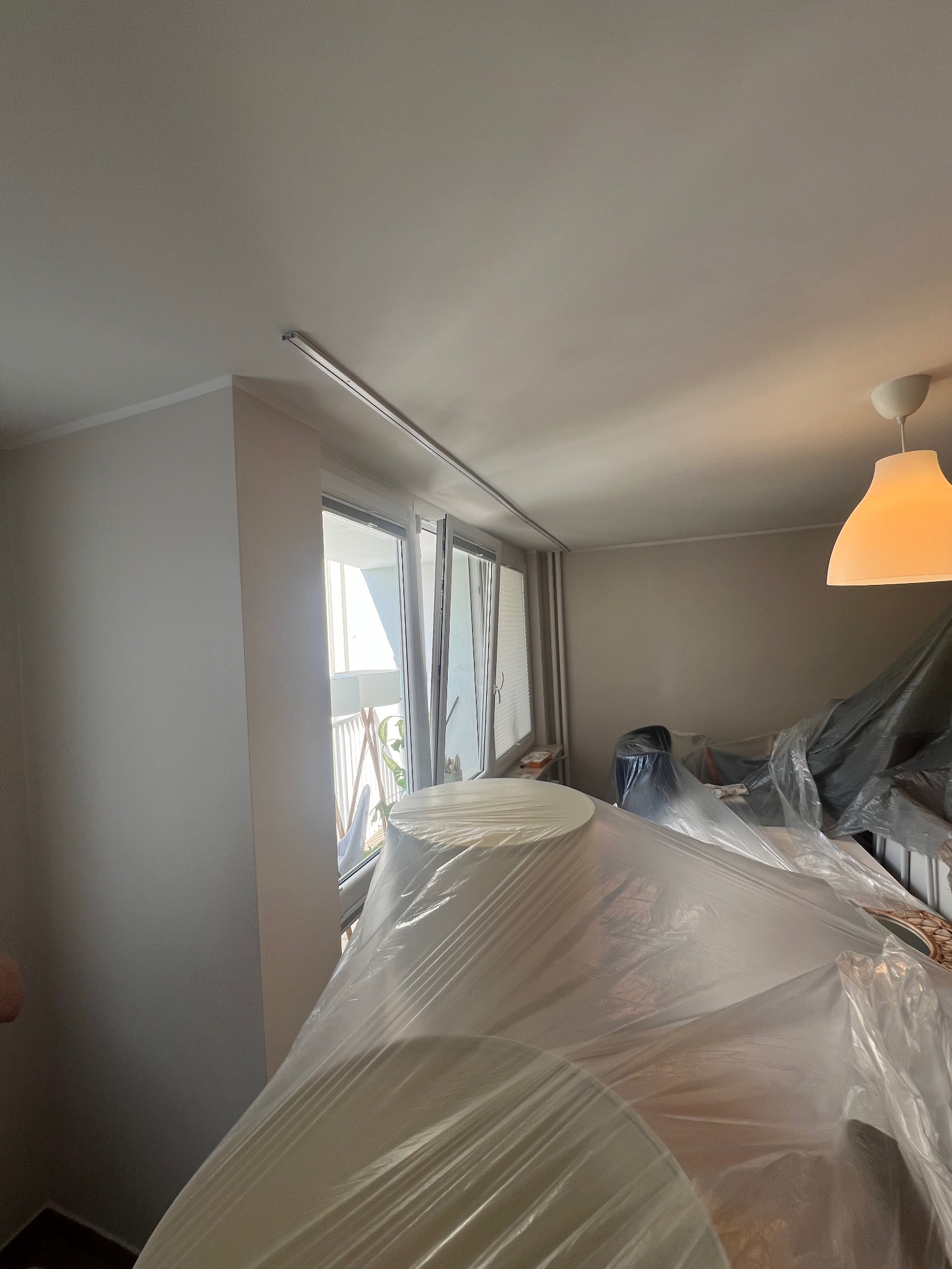 Pokój w trakcie prac remontowych: ściany po malowaniu, meble zabezpieczone folią malarską, widok na okno balkonowe z roletami. Przygotowanie do dalszych prac.