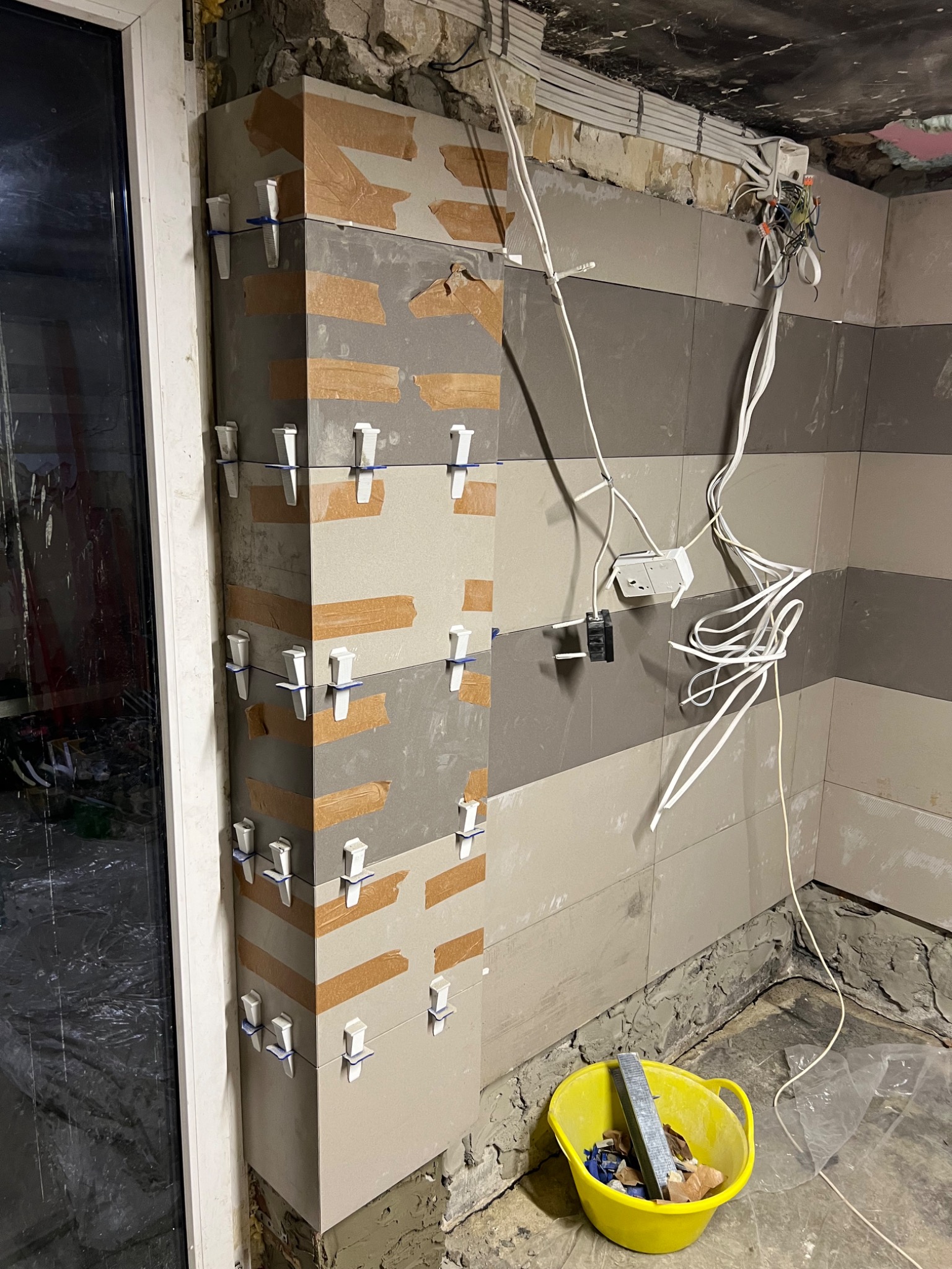 Układanie płytek na ścianie z użyciem klipsów i taśmy, widoczne przewody elektryczne i żółte wiadro z narzędziami. Prace glazurnicze w trakcie realizacji.