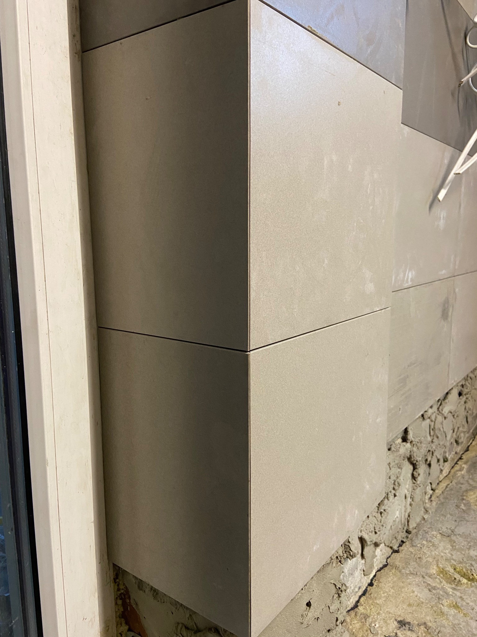 Wykończony narożnik ścian płytkami w odcieniach beżu i szarości. Widoczna dolna krawędź ściany przed położeniem płytek i fragment okna po lewej stronie.