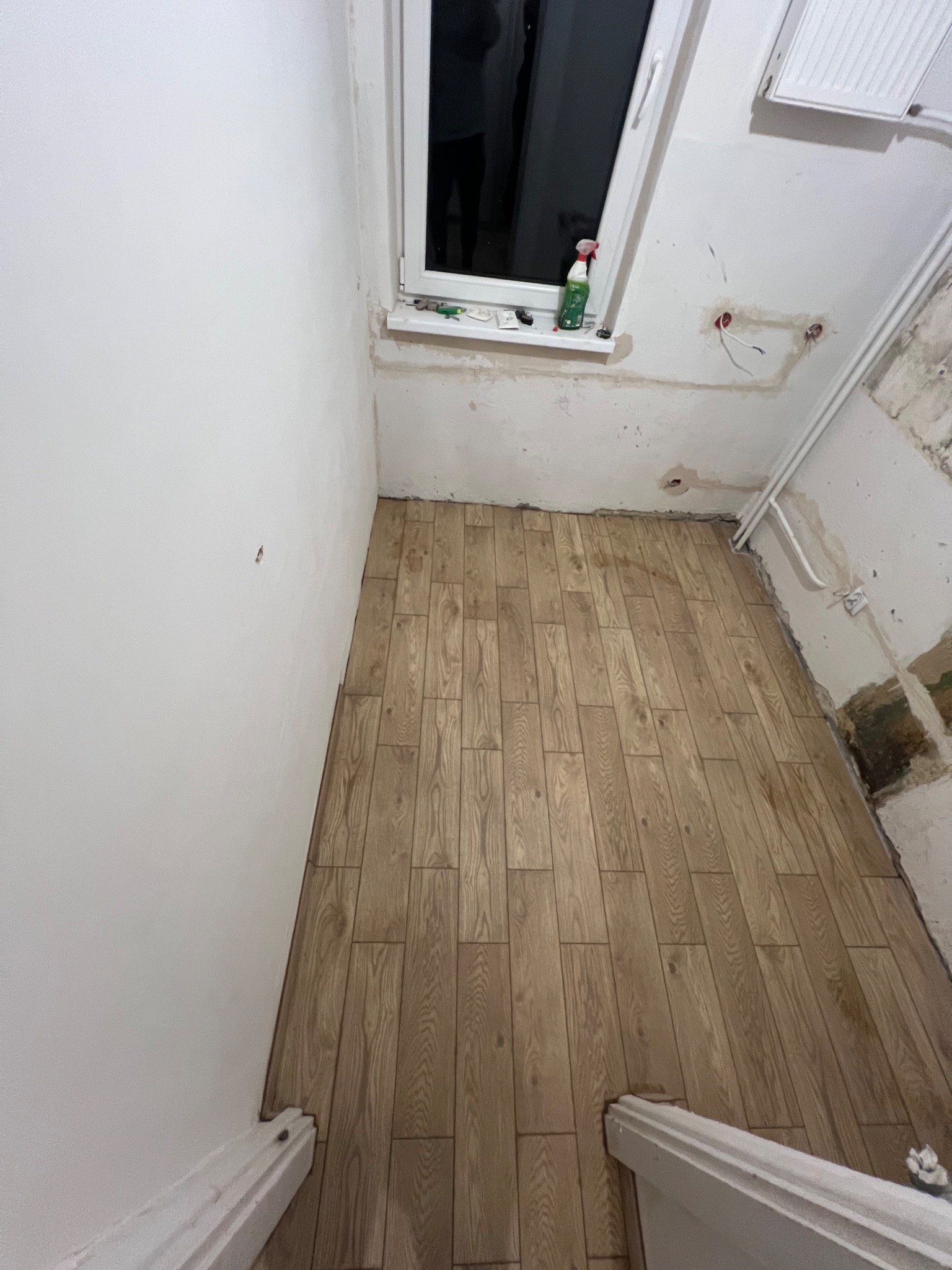 Ułożona podłoga z paneli imitujących drewno w trakcie remontu łazienki. Widoczne niedokończone ściany i instalacje. Na parapecie butelka ze środkiem czyszczącym.