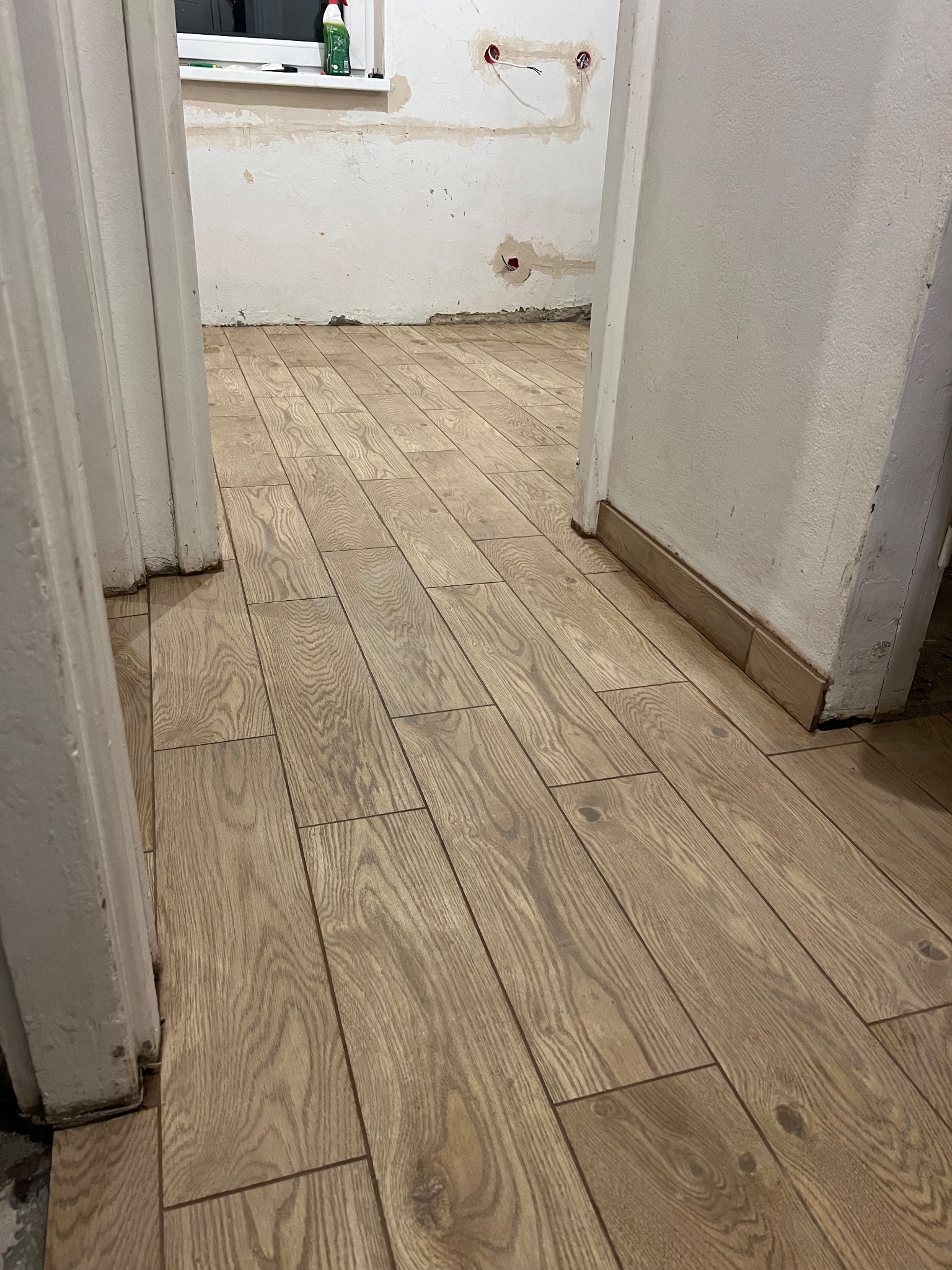 Ułożona podłoga z płytek imitujących drewno, widok z perspektywy wejścia do pomieszczenia. Białe futryny drzwiowe po bokach kadru, ściany w tle przygotowane do malowania.