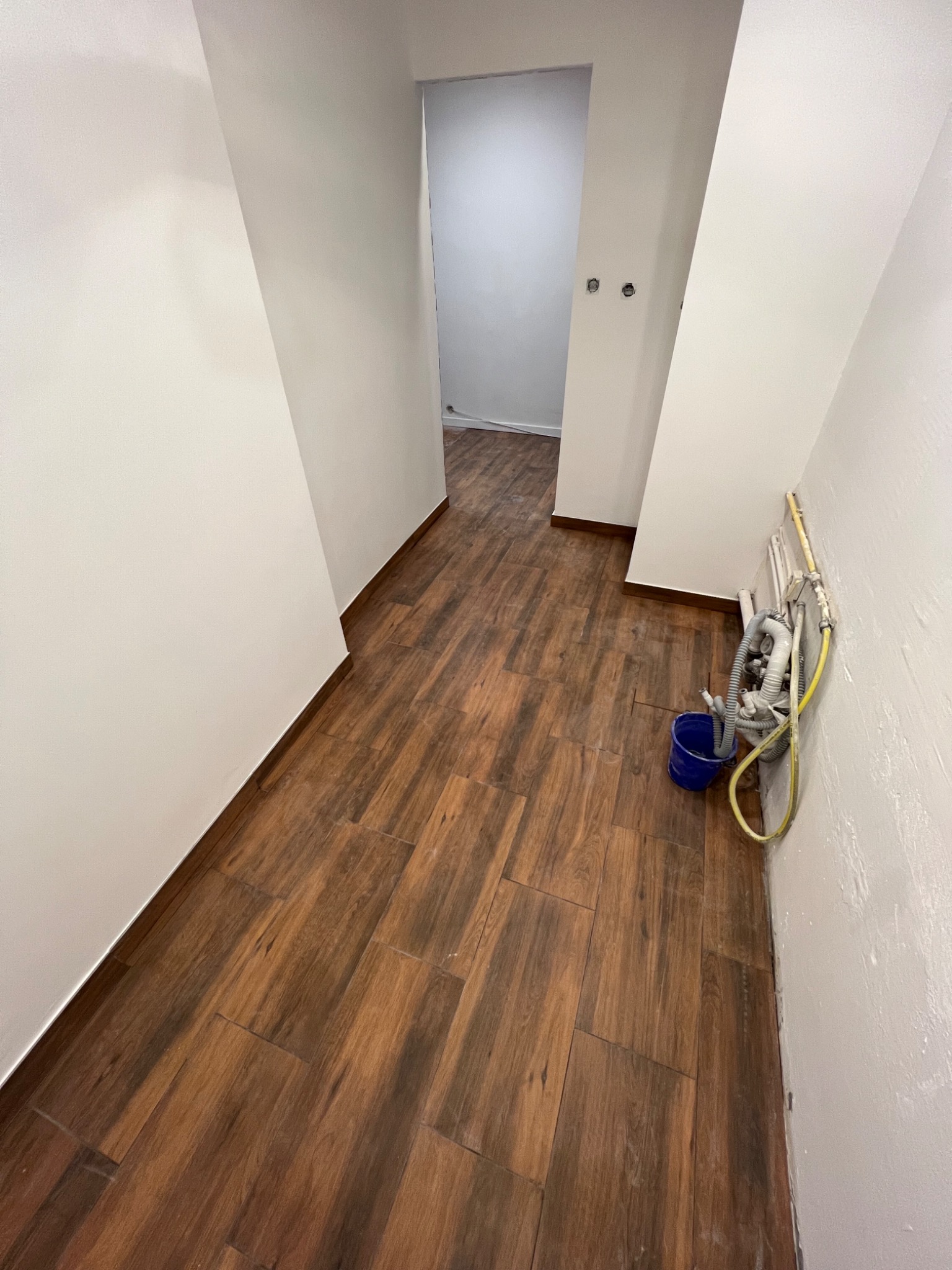 Korytarz po remoncie z brązową podłogą imitującą drewno. Na ścianie widoczne rury i niebieskie wiadro. Ściany białe, surowe, przygotowane do dalszych prac.