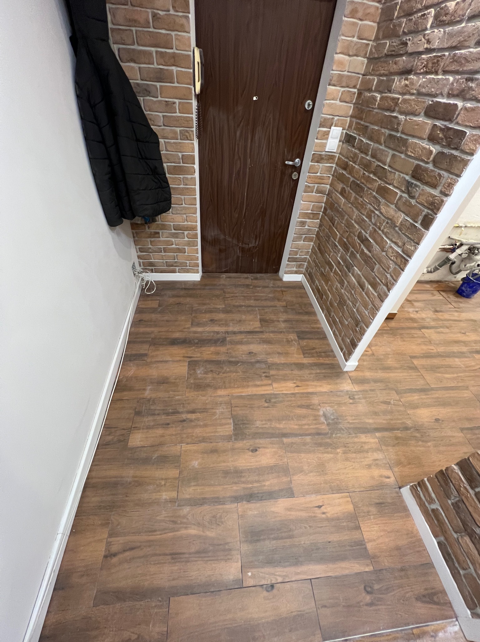 Korytarz po remoncie: brązowe płytki imitujące drewno na podłodze, ściana z cegły klinkierowej, ciemne drzwi wejściowe i czarna kurtka na wieszaku.