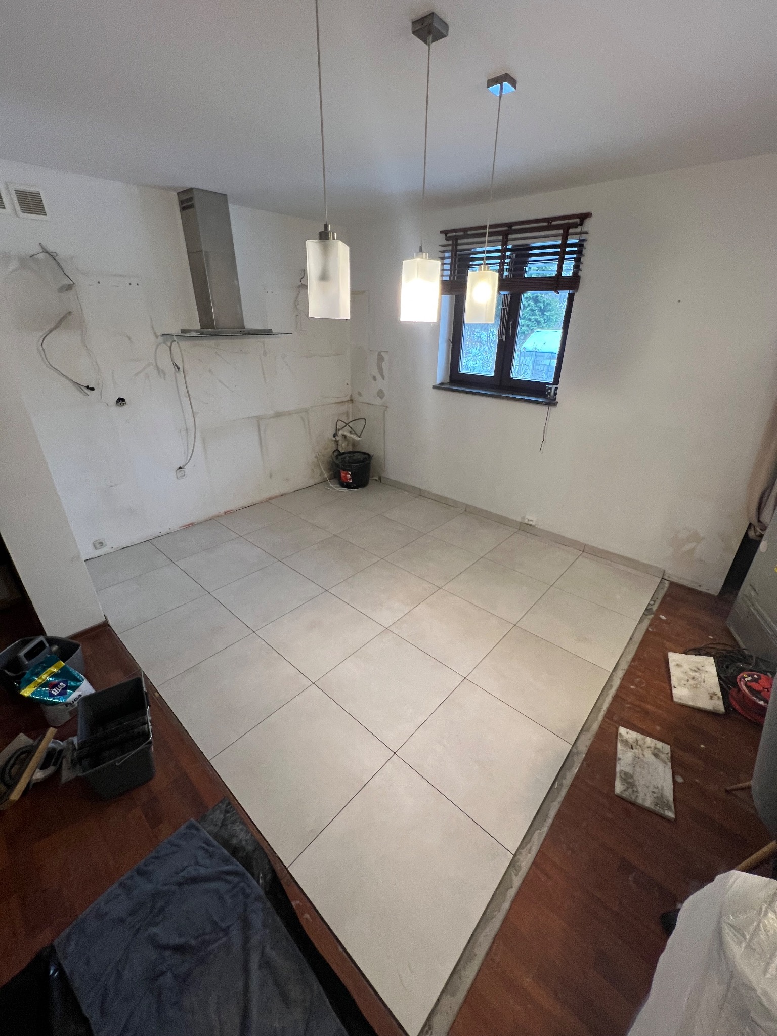 Ułożone jasne płytki podłogowe w trakcie remontu kuchni. Widoczne narzędzia i resztki materiałów budowlanych. Częściowo przygotowane ściany pod zabudowę.