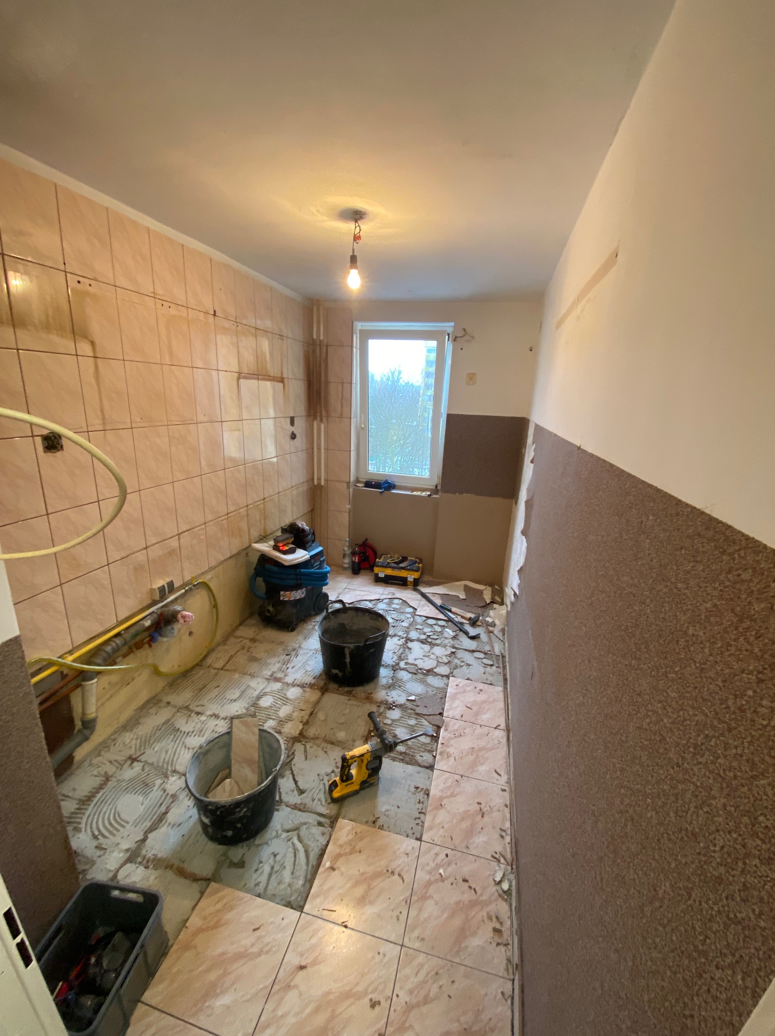 Łazienka w trakcie demontażu starych płytek. Widoczne narzędzia, wiaderka i pozostałości po kleju. Część ścian i podłogi bez okładzin.