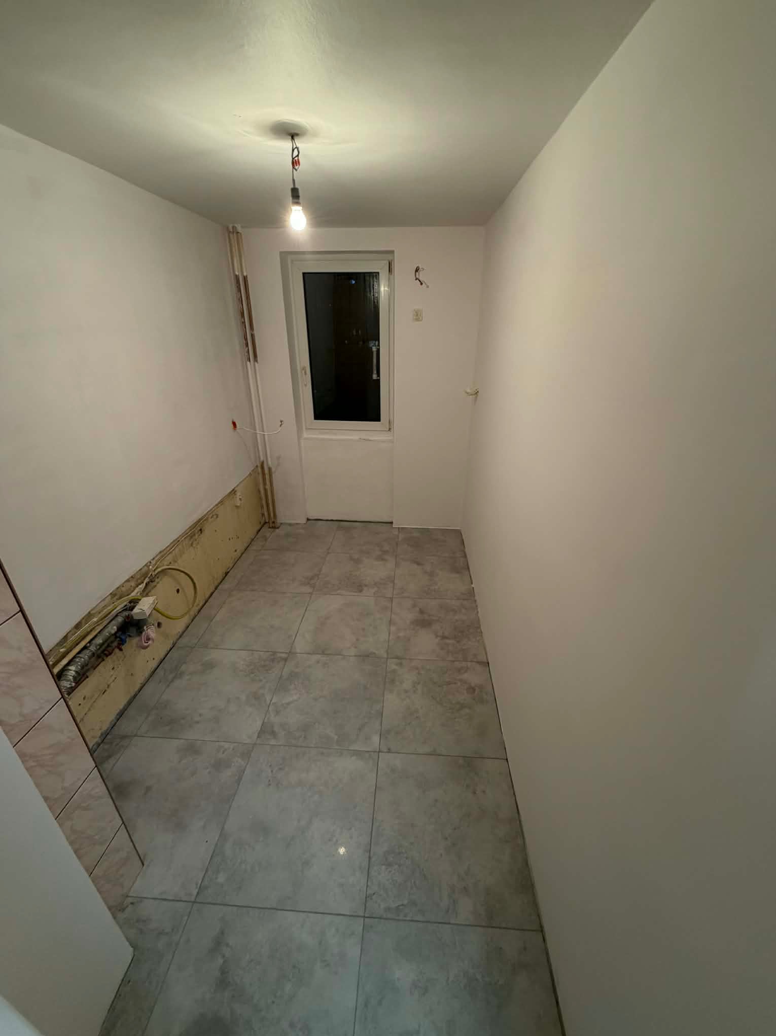 Wykończona podłoga z szarych płytek w małej łazience. Ściany częściowo przygotowane do malowania, widoczne instalacje. Żarówka na kablu oświetla wnętrze.