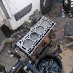 Rozłożony silnik samochodowy na stojaku warsztatowym, widoczne tłoki i głowica, obok biały samochód dostawczy i narzędzia. Układ cylindrów z bliska.