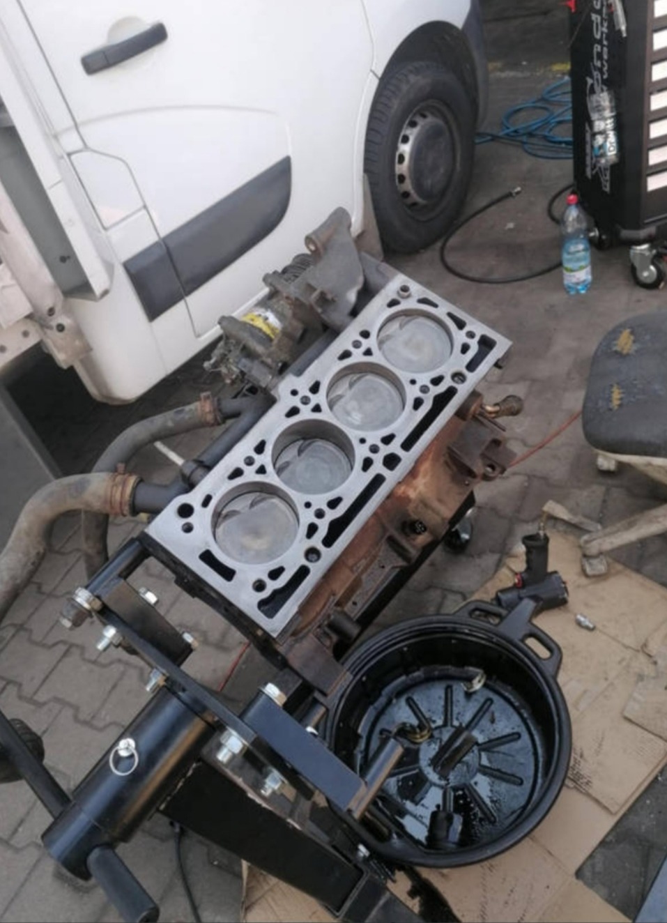 Rozłożony silnik samochodowy na stojaku warsztatowym, widoczne tłoki i głowica, obok biały samochód dostawczy i narzędzia. Układ cylindrów z bliska.