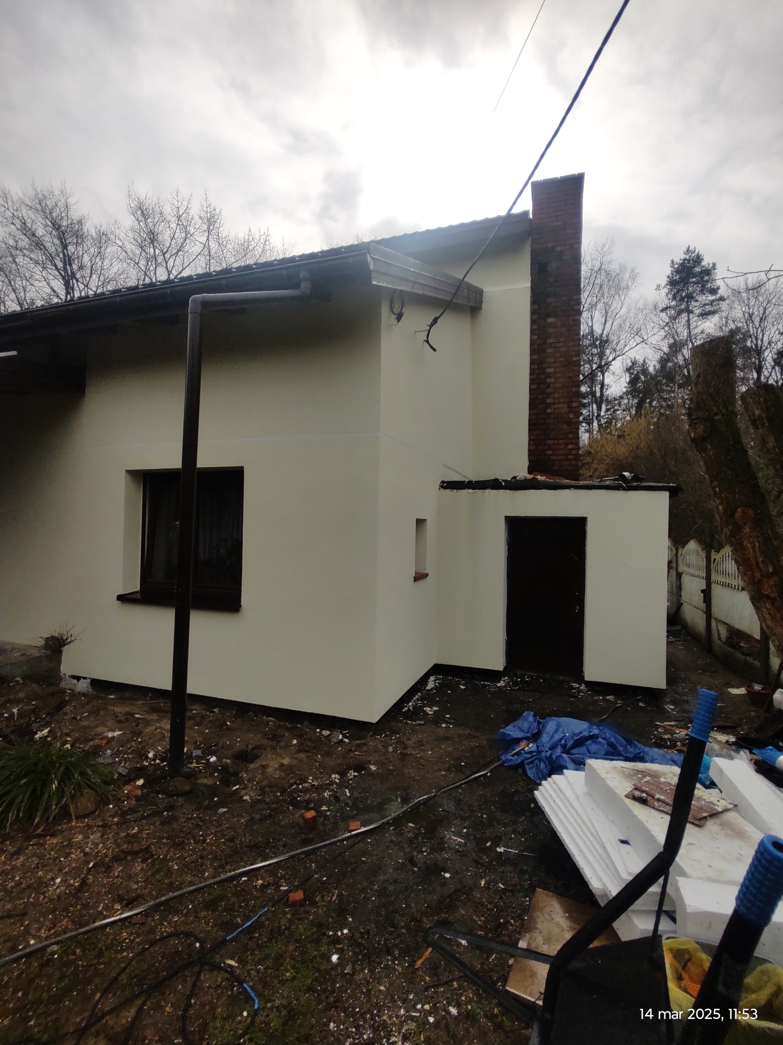 Świeżo pomalowana elewacja domu jednorodzinnego w jasnym odcieniu, widoczny ceglany komin, okno z ciemną ramą i przygotowane materiały budowlane na ziemi.