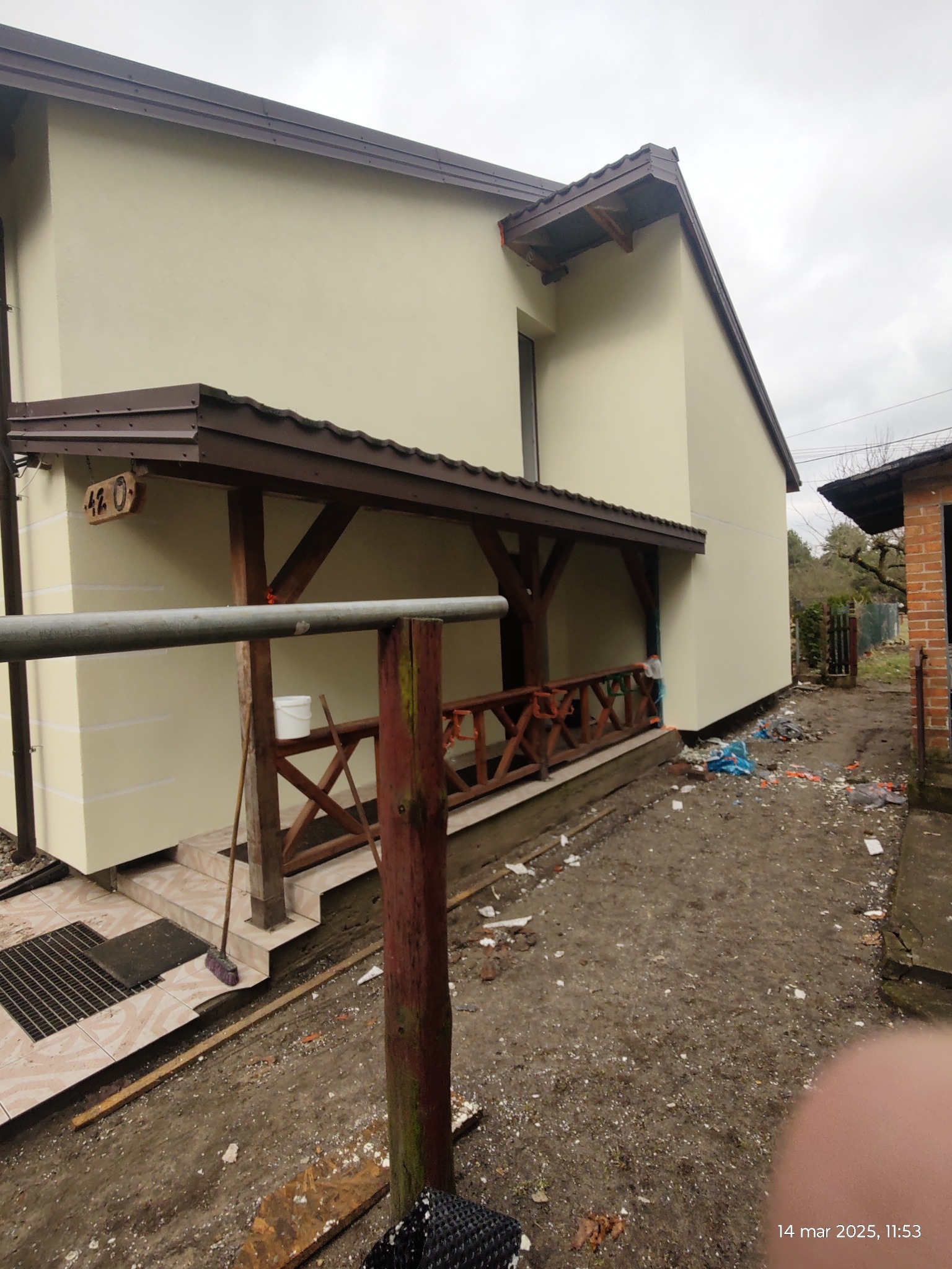 Świeżo otynkowana elewacja domu jednorodzinnego z drewnianym zadaszeniem nad wejściem, widoczne ślady prac budowlanych na ziemi, numer domu '420' na elewacji.