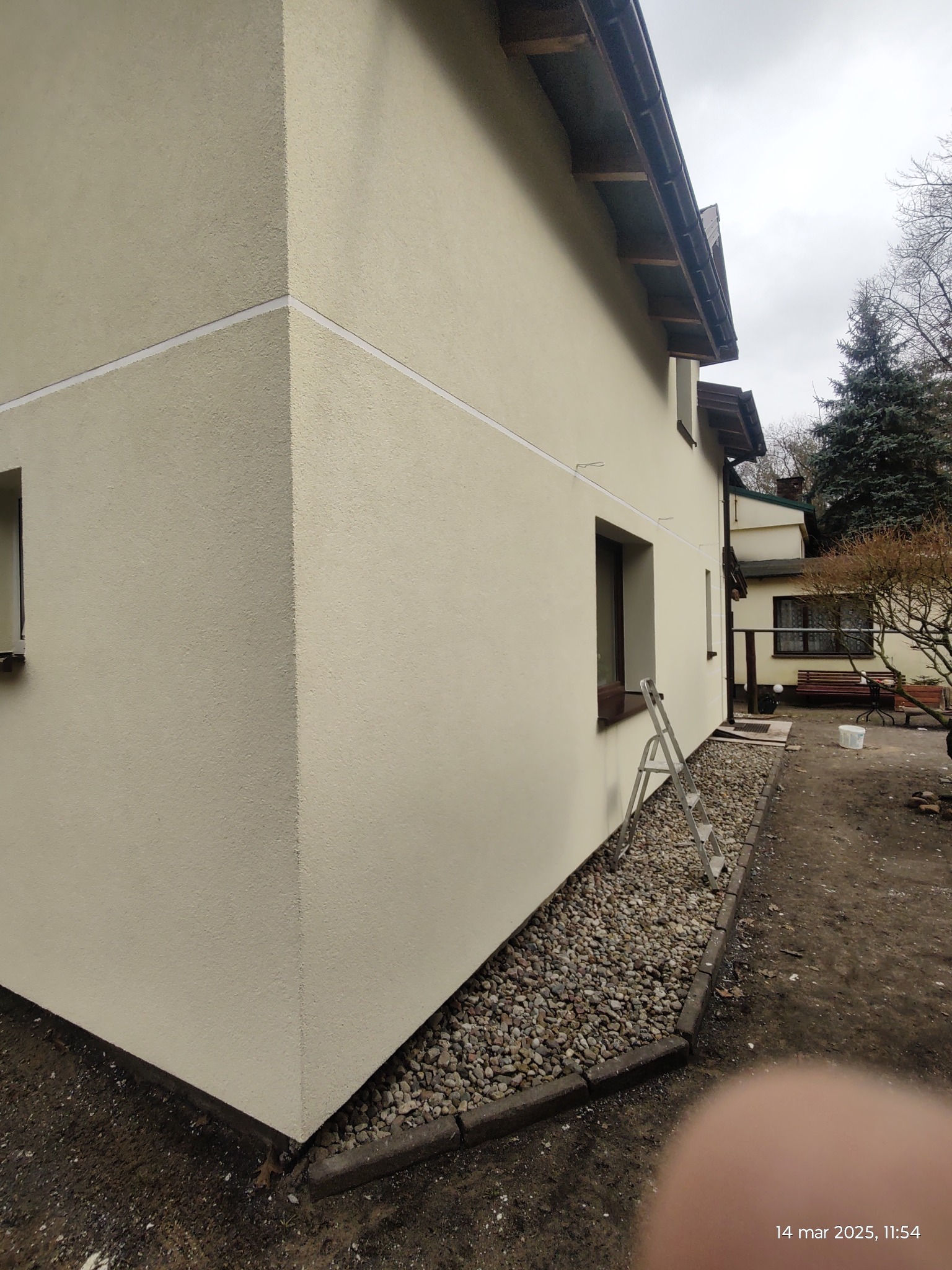 Jasna elewacja domu jednorodzinnego po tynkowaniu, z widoczną listwą dekoracyjną i drabiną opartą o ścianę, widok z boku w pochmurny dzień.