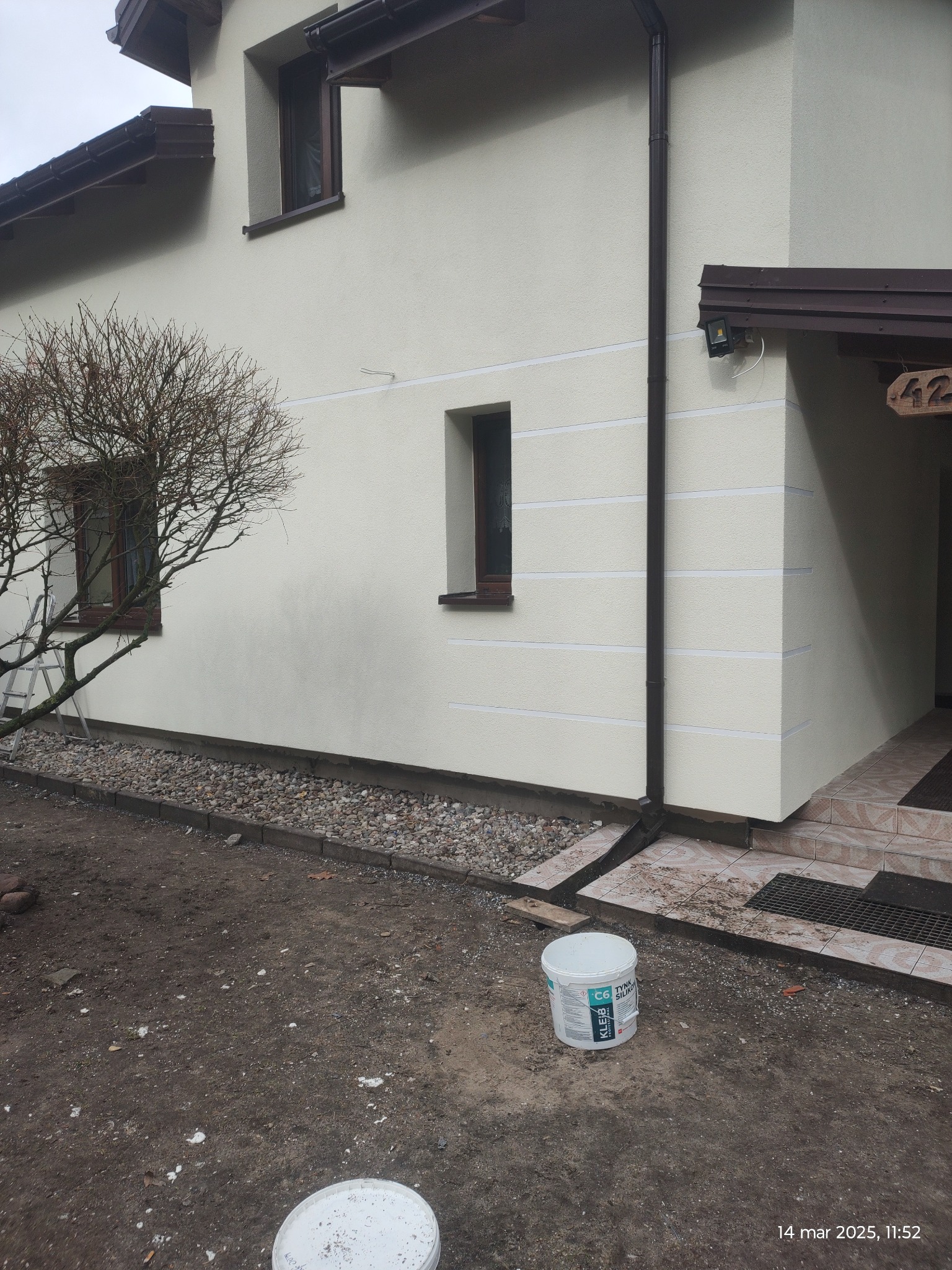 Ocieplony budynek z jasną elewacją, z widocznymi poziomymi liniami dekoracyjnymi, brązowymi oknami i rynnami, oraz wiadro z tynkiem silikonowym C6 na pierwszym planie.