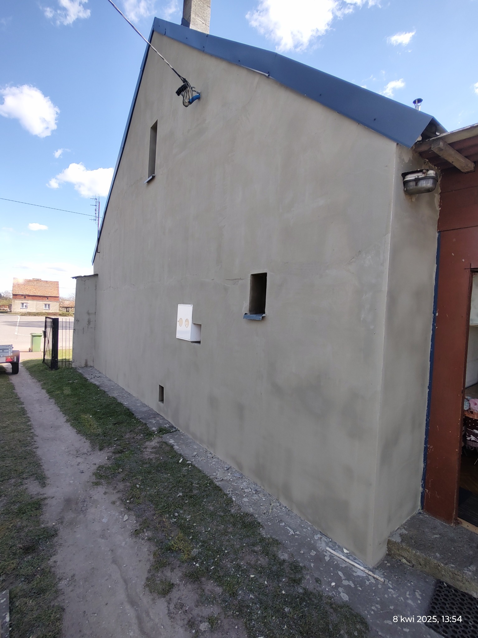 Ściana budynku po tynkowaniu elewacji, widoczne okna bez oprawy, skrzynka elektryczna i fragment dachu z niebieską blachą.