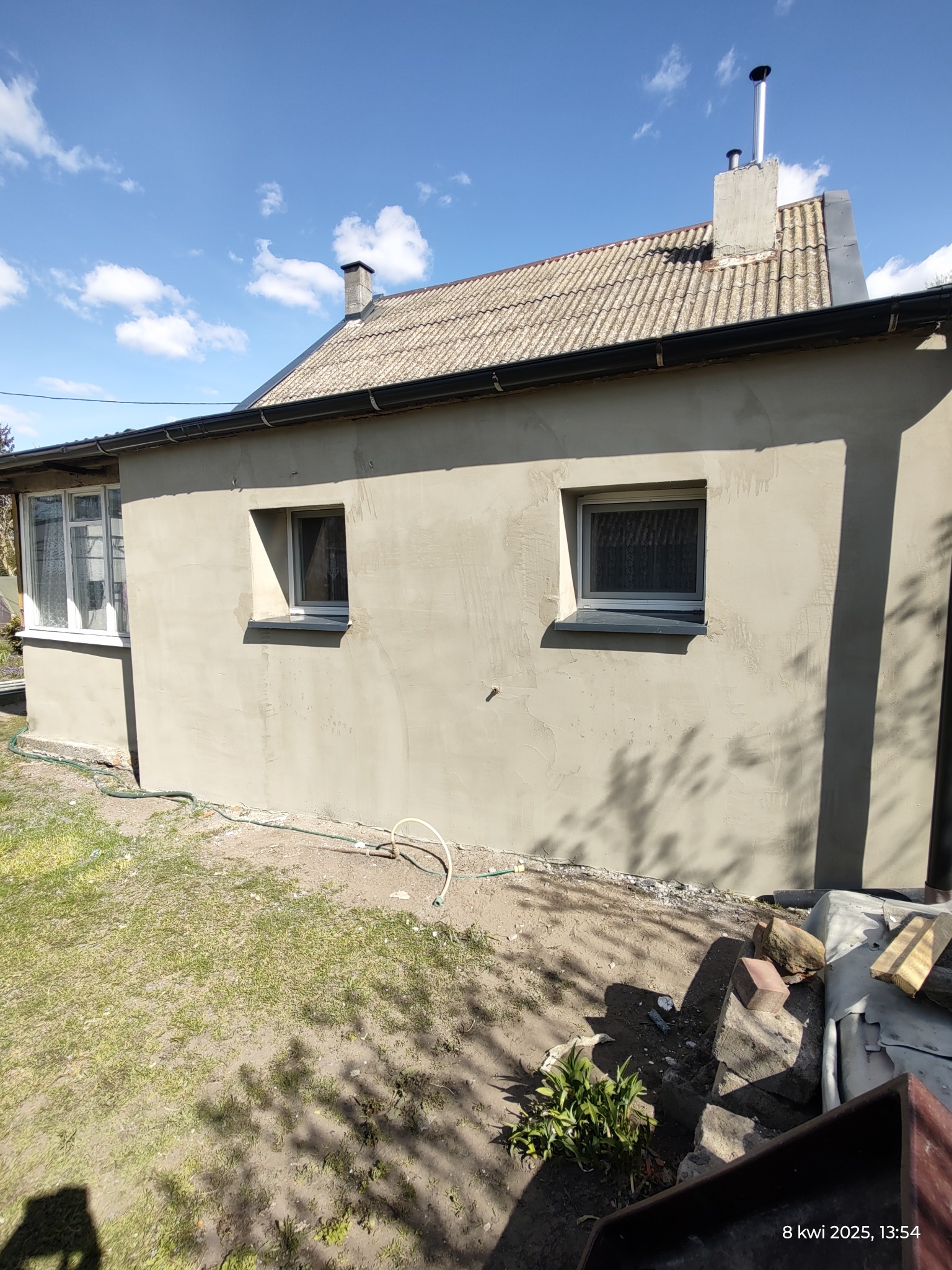 Świeżo otynkowana elewacja domu jednorodzinnego z dwoma oknami, widoczne nierówności tynku i przygotowane podłoże wokół budynku.