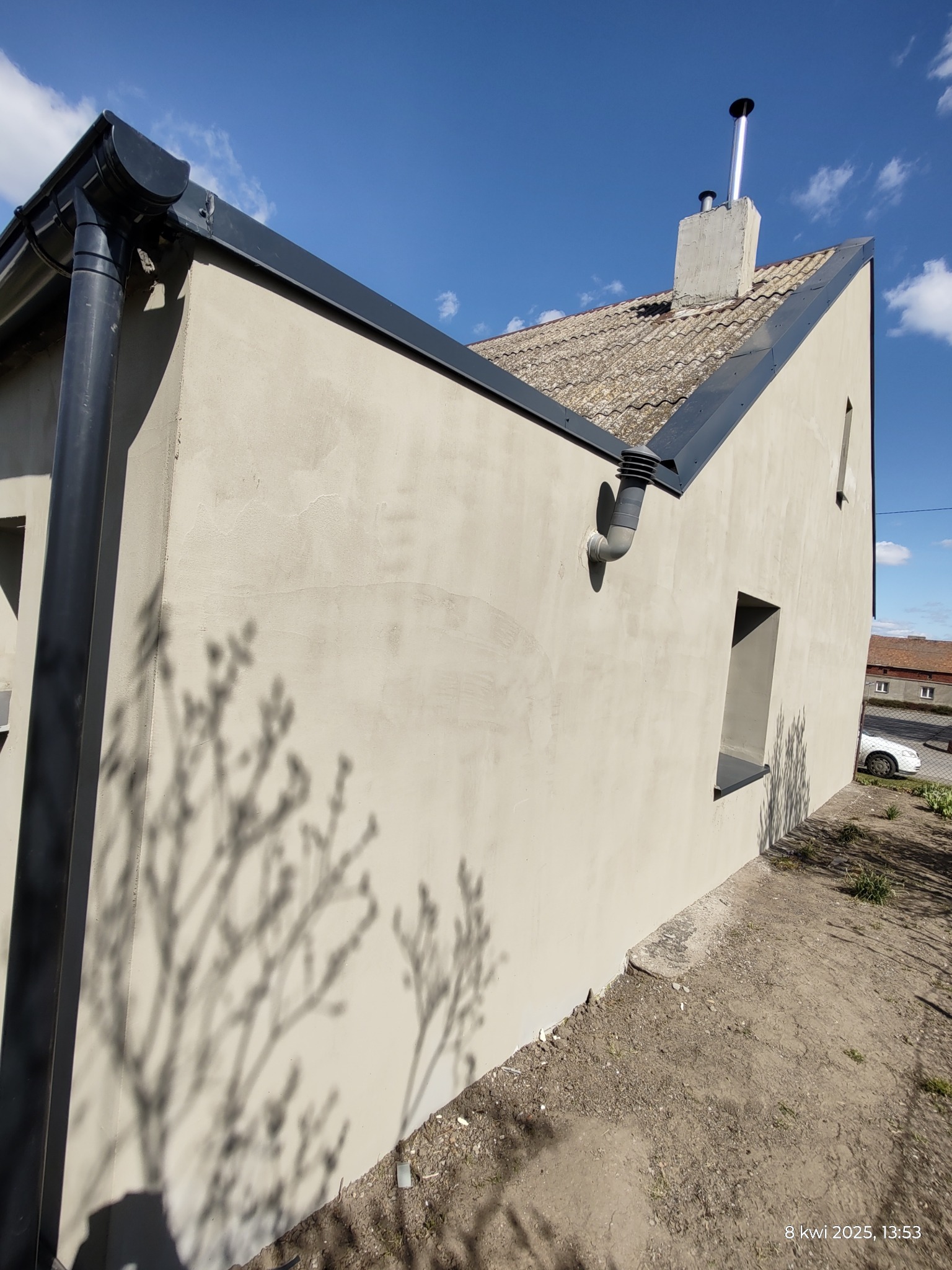 Świeżo otynkowana, jasna elewacja domu jednorodzinnego z widocznym kominem i systemem rynnowym, cień gałęzi na ścianie, w tle błękitne niebo.