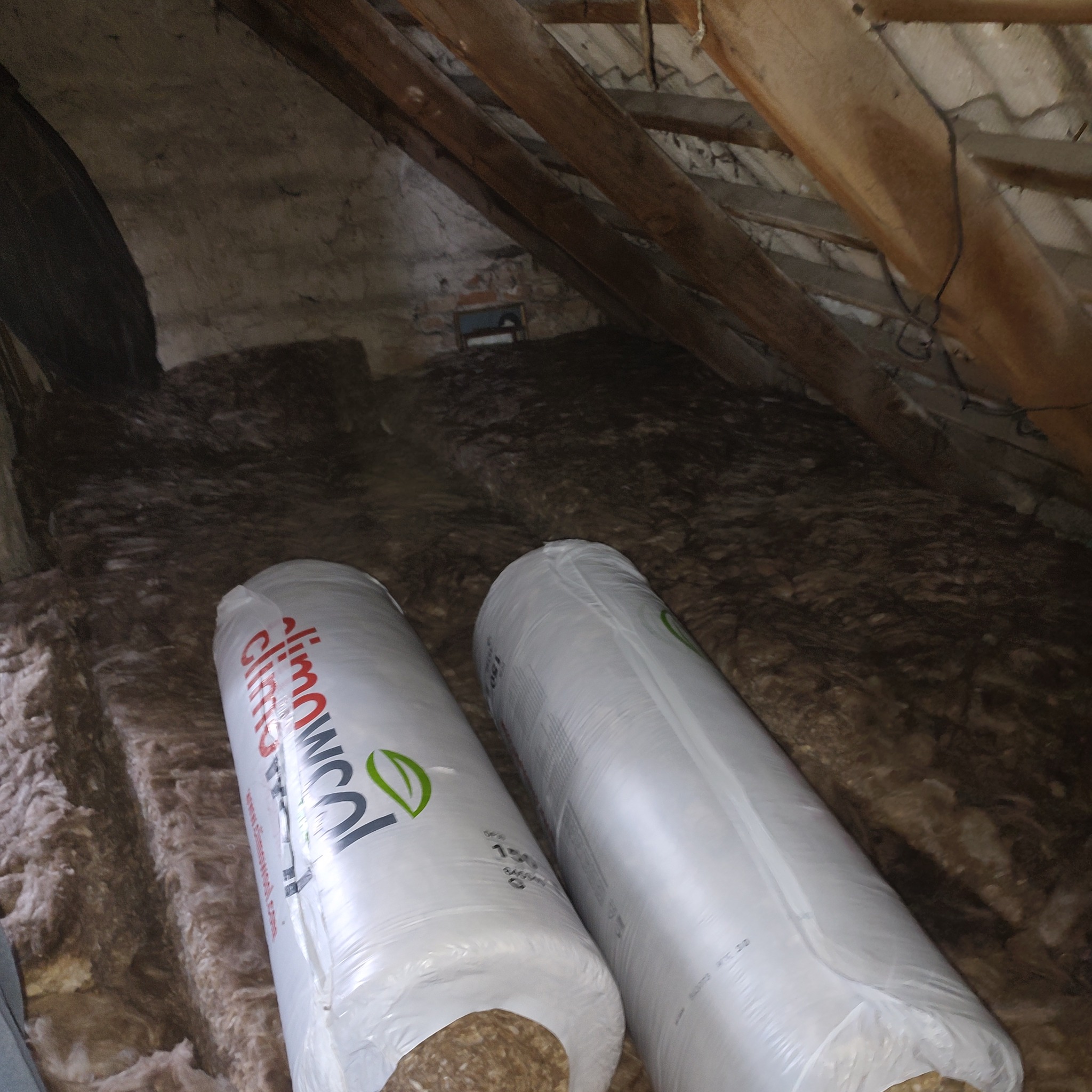 Dwa zrolowane opakowania wełny mineralnej Climowool leżą na poddaszu z widocznymi drewnianymi belkami stropowymi i fragmentami starej ściany.