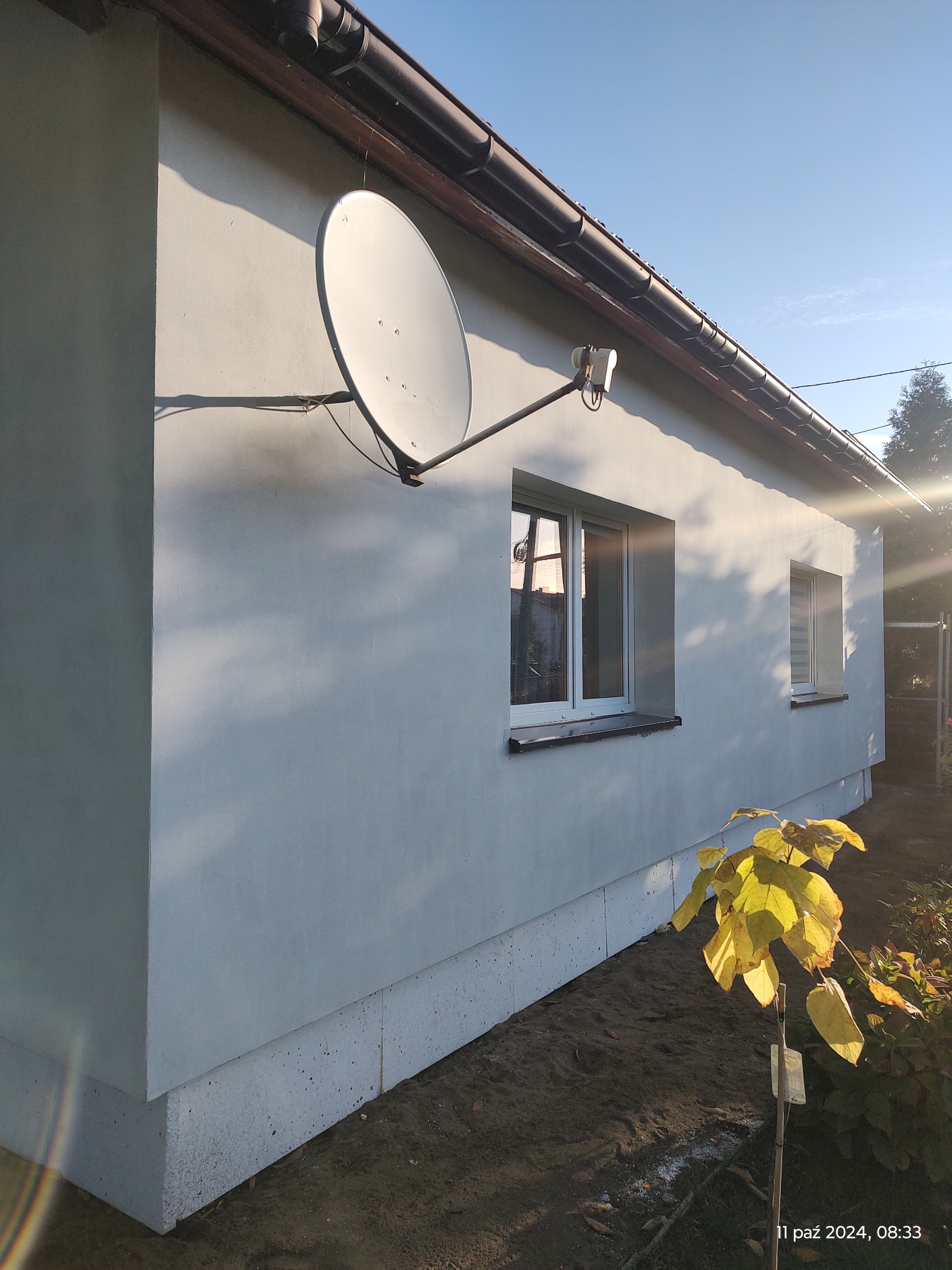 Otynkowana elewacja domu jednorodzinnego z anteną satelitarną, widoczne okna i świeżo obsypana ziemia wokół budynku.