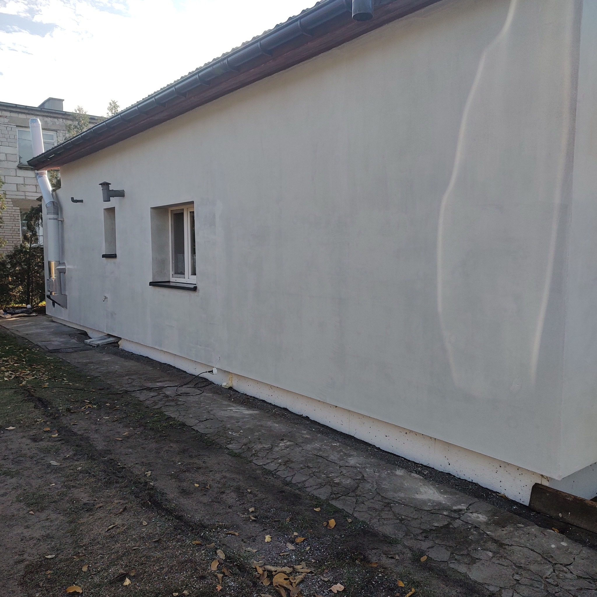 Świeżo otynkowana i pomalowana elewacja budynku z widocznymi oknami, rynnami i kominem, otoczona jesiennymi liśćmi na ziemi.