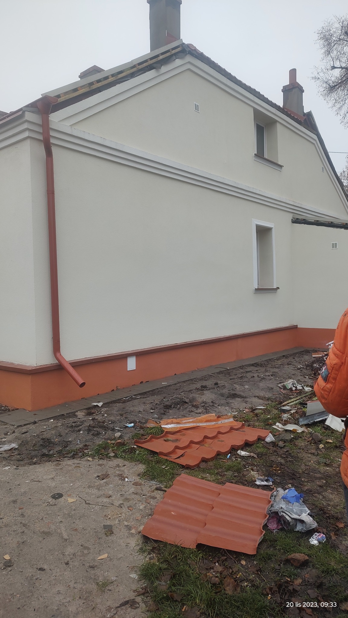 Świeżo otynkowana i pomalowana elewacja domu jednorodzinnego z widocznymi detalami architektonicznymi, takimi jak listwy ozdobne i cokół w kolorze ceglastym, z fragmentami dachu i rynny,...