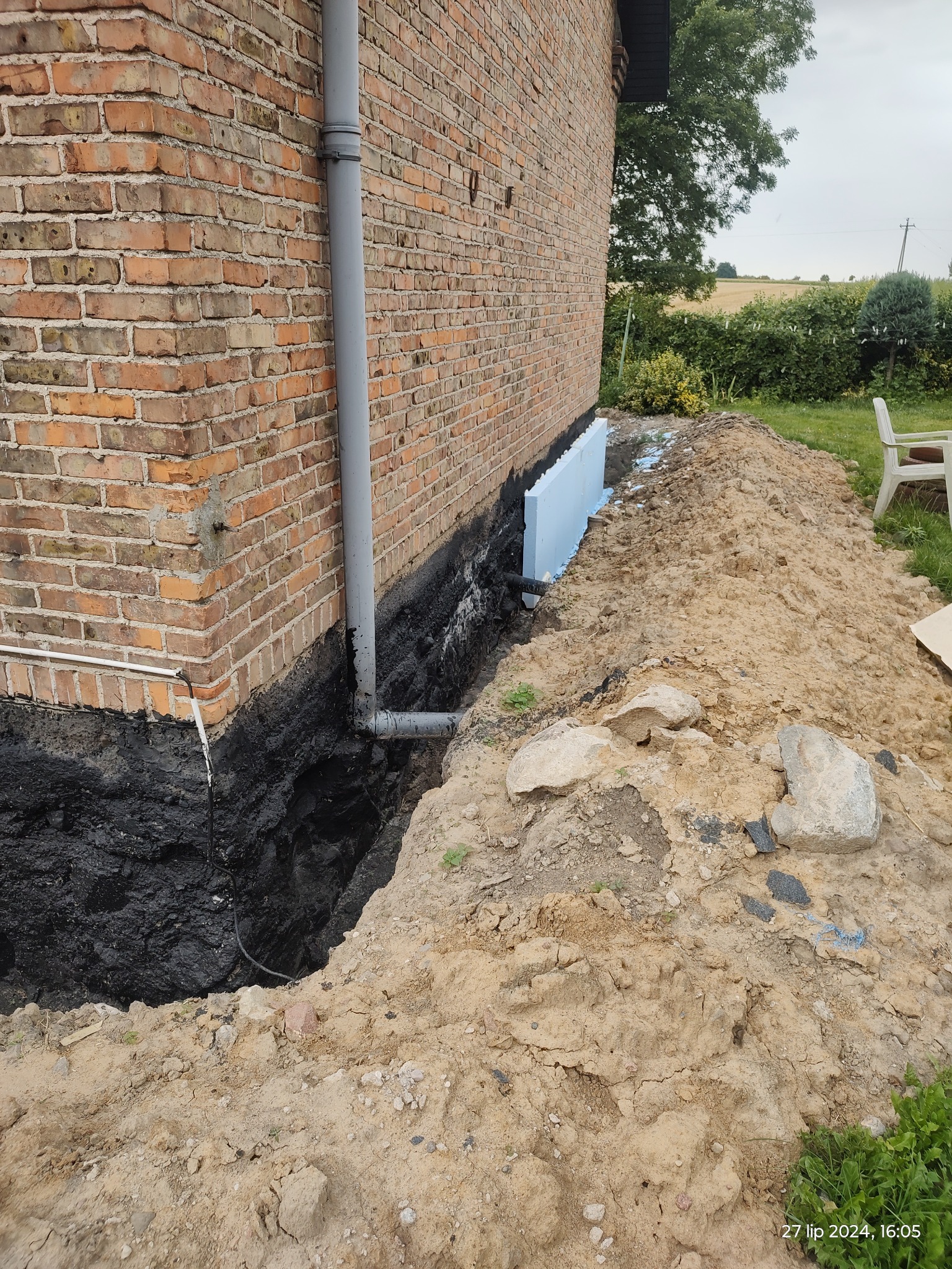 Ceglana ściana budynku z widoczną izolacją fundamentów, rurą spustową i wykopem zasypanym ziemią.