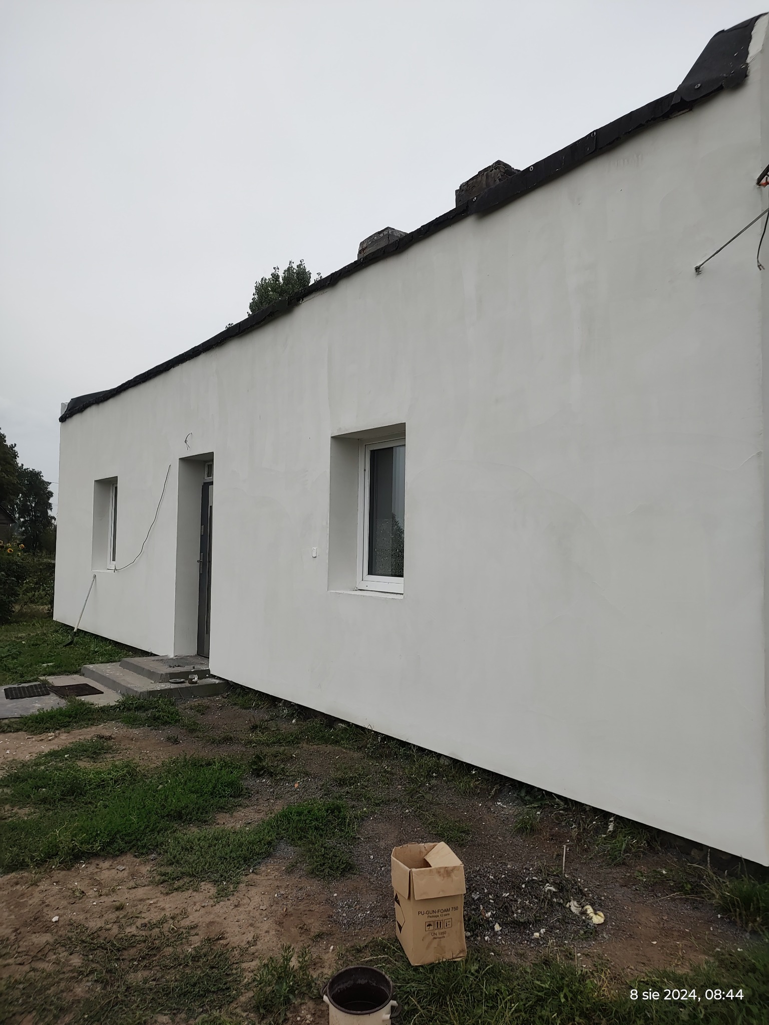 Świeżo otynkowana, biała elewacja domu jednorodzinnego z widocznymi śladami po tynkowaniu, karton po piance montażowej leżący na ziemi, pochmurne niebo.