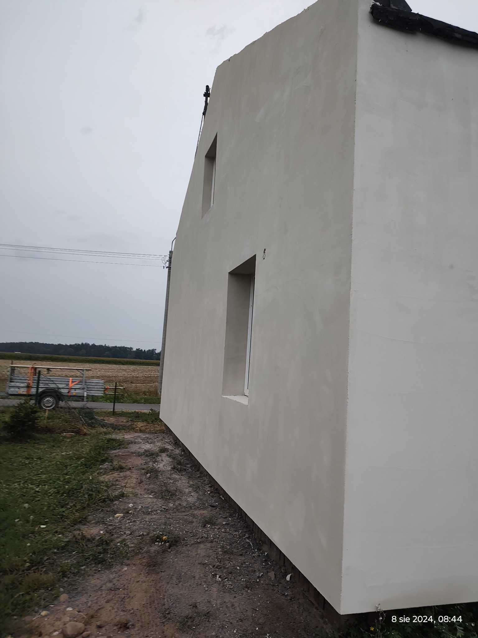 Świeżo otynkowana ściana domu jednorodzinnego z dwoma oknami, widok z boku, na tle pochmurnego nieba i pola, z przyczepką transportową w tle.