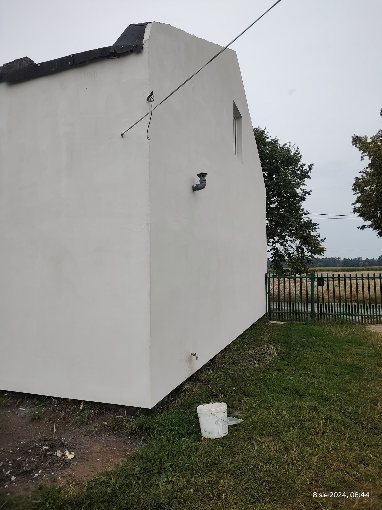 Świeżo otynkowana ściana domu z widocznym kominkiem wentylacyjnym, fragmentem okna i wiadrem po farbie leżącym na trawie.