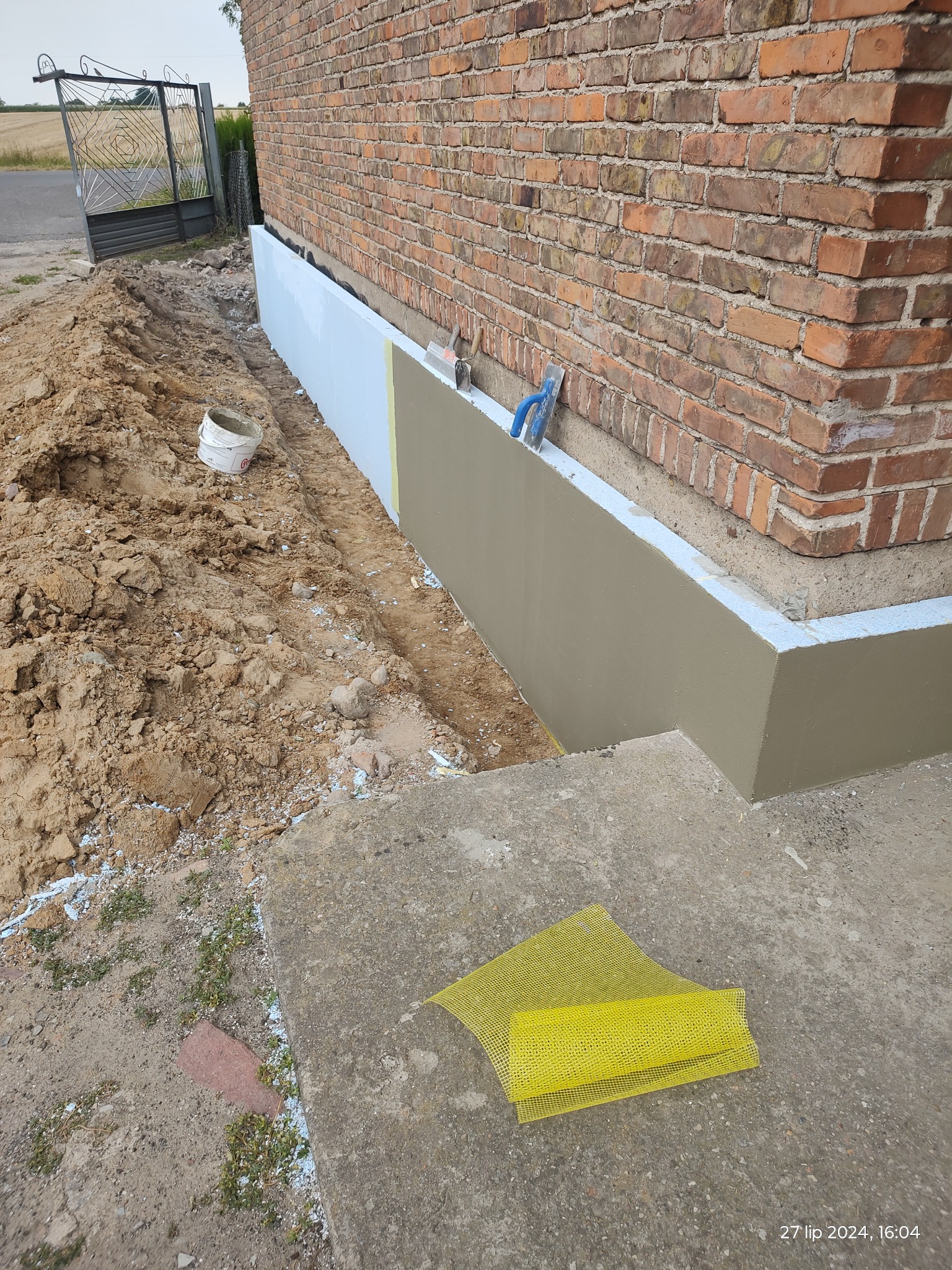 Częściowo ocieplona ściana fundamentowa ceglanego budynku z widoczną warstwą styropianu, siatką elewacyjną i narzędziami murarskimi.