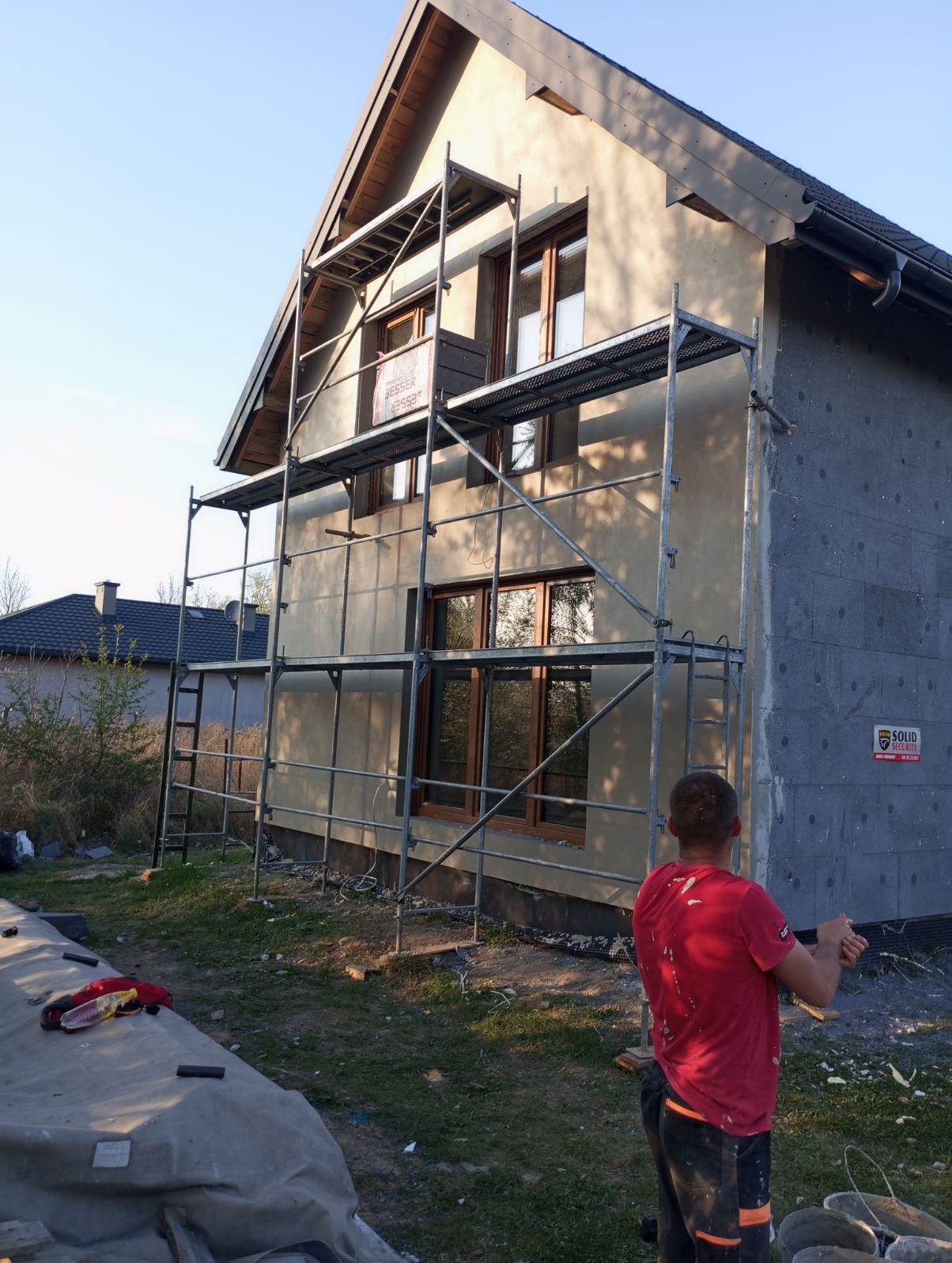 Dom jednorodzinny w trakcie ocieplania, widoczne rusztowanie, płyty izolacyjne na ścianie szczytowej i fragment nałożonego tynku na pozostałej części elewacji.