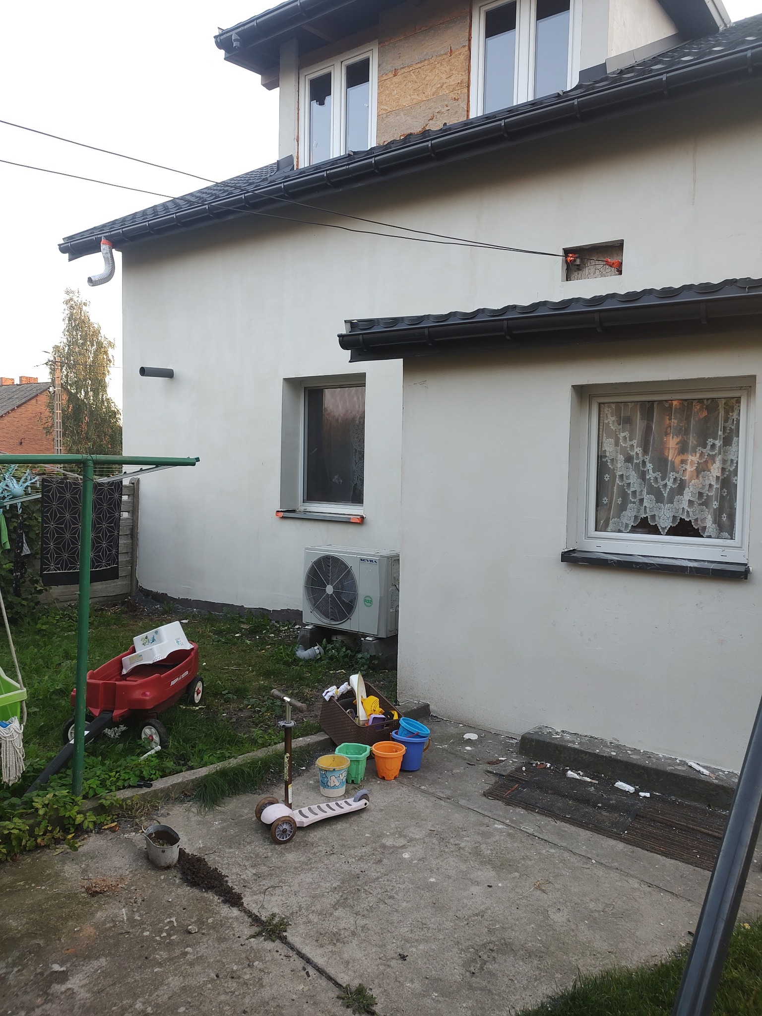 Ściana domu po tynkowaniu na biało, z widocznym otworem na kabel, oknami z białymi ramami i klimatyzatorem, na zewnątrz zabawki dziecięce i pranie.