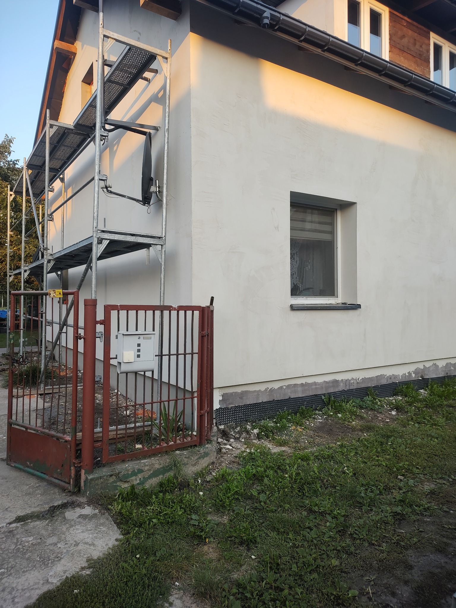 Dom jednorodzinny w trakcie tynkowania elewacji, widoczne rusztowanie i fragment ogrodzenia z bramką.