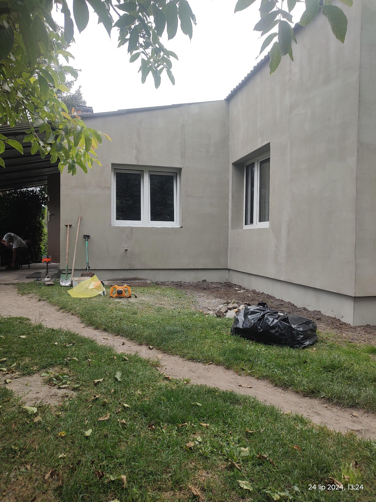 Dom po tynkowaniu elewacji, widoczne narzędzia, worki z odpadami i pracownik w tle.