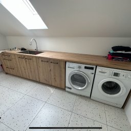 Bartosz Stokłosa - Pralka i suszarka zabudowane pod blatem z drewnianym wykończeniem, zintegrowane z szafkami i zlewem w pomieszczeniu gospodarczym na poddaszu ze świetlikiem.