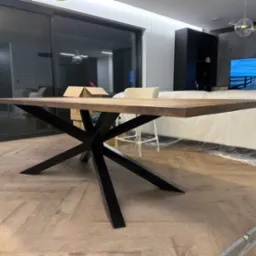 Stół z blatem z ciemnego drewna i czarną, geometryczną podstawą w nowoczesnym wnętrzu z dużym oknem przesuwnym i drewnianą podłogą w jodełkę. W tle widoczna sofa zabezpieczona folią malarską.